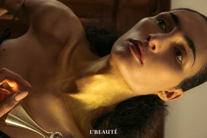 Adult female lying down with a golden, glossy skin tone. She is holding a perfume bottle in soft, diffused lighting. Beauty shot.
