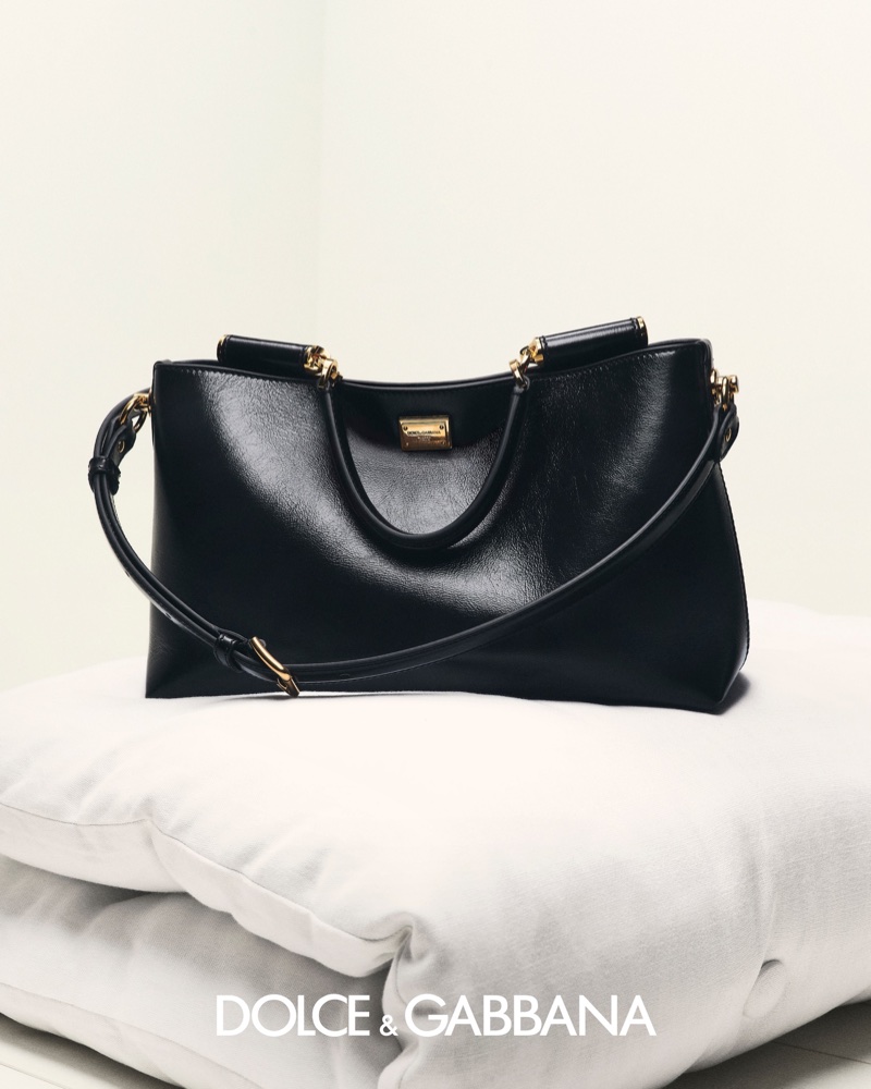 Black Dolce & Gabbana leather handbag with gold hardware resting on a white cushion against a pale studio background.