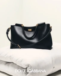 Black leather handbag with gold hardware resting on white cushions against a plain white studio background.