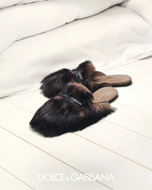 Fluffy brown fur slip-on mules with brand labels resting on a white wooden floor in front of white bedding.
