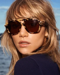 Close-up portrait of a woman wearing tortoiseshell aviator sunglasses and a dark blue ribbed top against an outdoor water background.