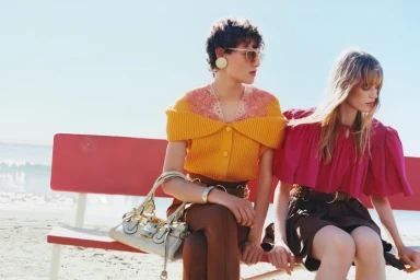 Two women seated outdoors on a red beach bench wearing colourful blouses and holding a designer handbag against the ocean horizon