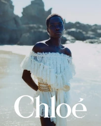 Model wearing an off-the-shoulder white lace dress with a metallic belt standing by the rocky coast.