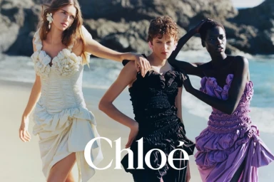 Three female models posing in ruffled dresses on a sandy beach against rocky coastlines for Chloe Spring-Summer 2026 campaign.