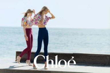Two female models posing in floral crop tops and high waisted trousers on a wooden platform overlooking the ocean under a clear sky.