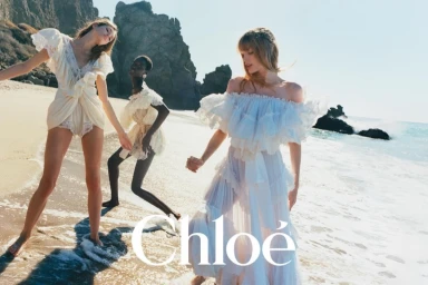 Three models wearing flowing white and cream dresses on a sandy beach near rocky outcrops with ocean waves breaking.