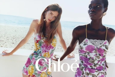 Two models in floral print swimwear leaning on a white wall overlooking the beach for the Chloé Spring-Summer 2026 campaign.