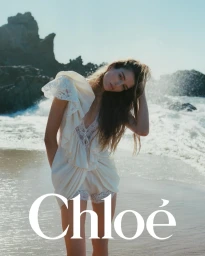 Model wearing a white lace dress standing on a sandy beach near ocean waves under clear sky