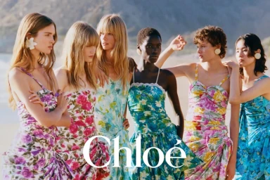 Group of six female models wearing floral dresses standing on a sandy beach for Chloe Spring-Summer 2026 campaign.