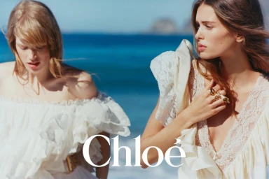 Two female models in white lace dresses posing outdoors against a blue ocean background for Chloé.