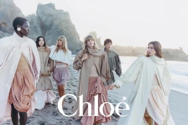 Group of six female models wearing neutral-toned layered coats and skirts posing on a sandy beach at sunset for Chloé.