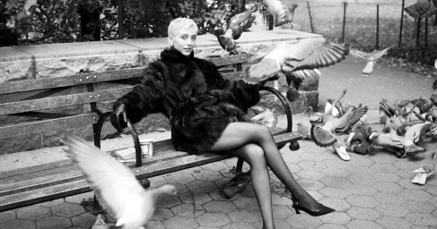 Emma Chamberlain in a black fur coat sitting on a park bench surrounded by flying pigeons for Stuart Weitzman