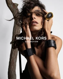 Model Mica Arganaraz wearing black leather cuffs and a large gold ring with tousled dark hair against a plain white background.