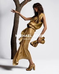 Model Mica Arganaraz in a metallic gold dress holding a matching clutch against a white seamless backdrop with a tree trunk prop.