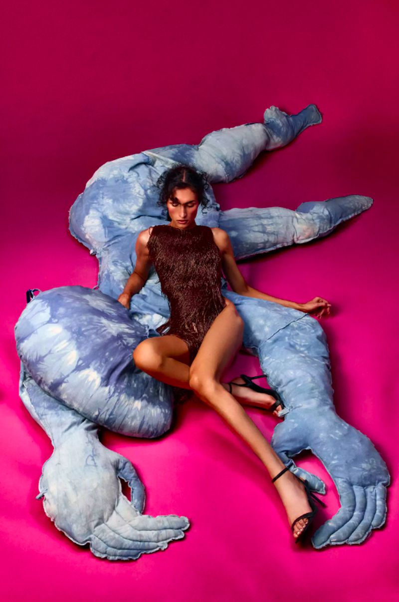 Woman in a brown leotard poses on a blue and white abstract sculpture against a vivid pink backdrop in a studio setting.