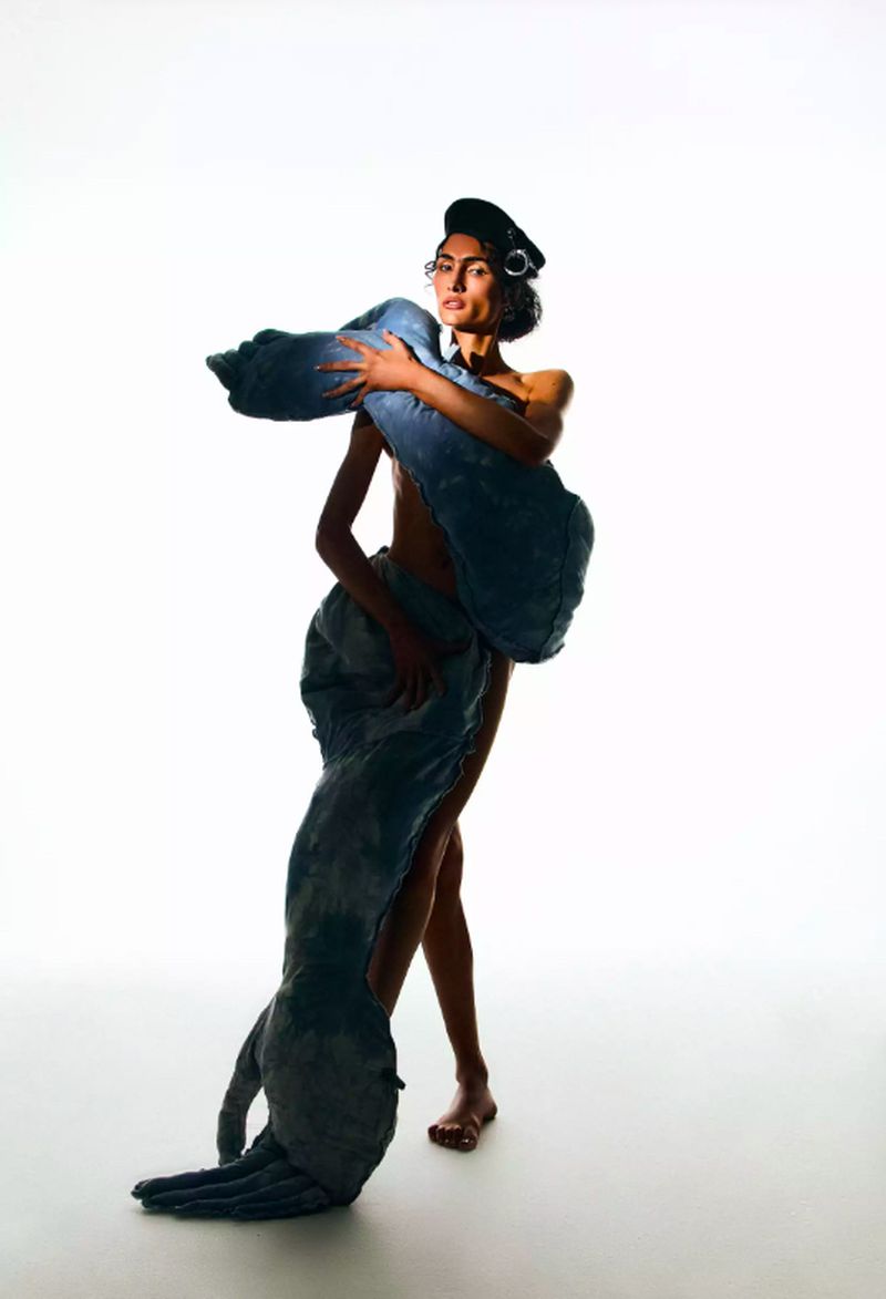 An adult female poses in a photo studio wearing a black beret and a large, sculpted denim textile dress. The background is white.