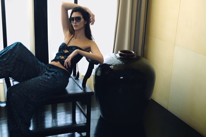Vittoria Ceretti in dark sunglasses and a shimmering petrol blue two-piece set lounging on a dark chair next to a large ceramic vase.