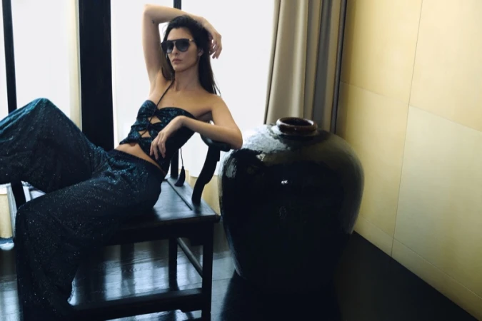Vittoria Ceretti in dark sunglasses and a shimmering petrol blue two-piece set lounging on a dark chair next to a large ceramic vase.