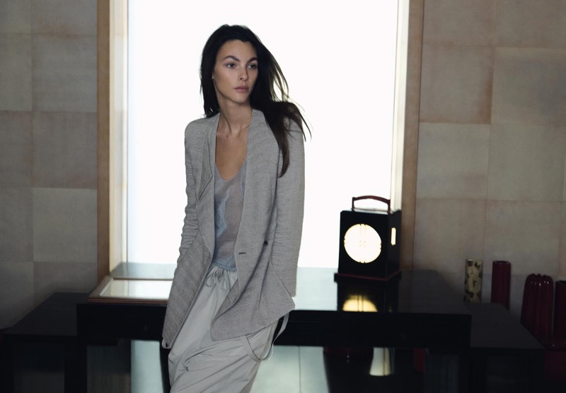 Model Vittoria Ceretti wearing a grey jacket and light trousers leaning against a dark table for Giorgio Armani
