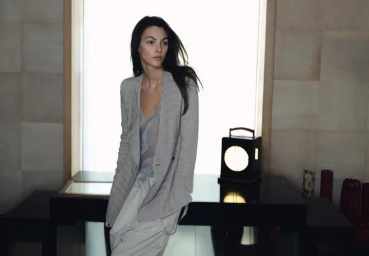 Model Vittoria Ceretti wearing a grey jacket and light trousers leaning against a dark table for Giorgio Armani
