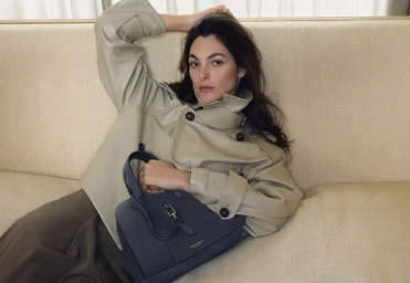 Vittoria Ceretti reclining on a cream sofa wearing a light tan leather jacket and holding a dark grey handbag.