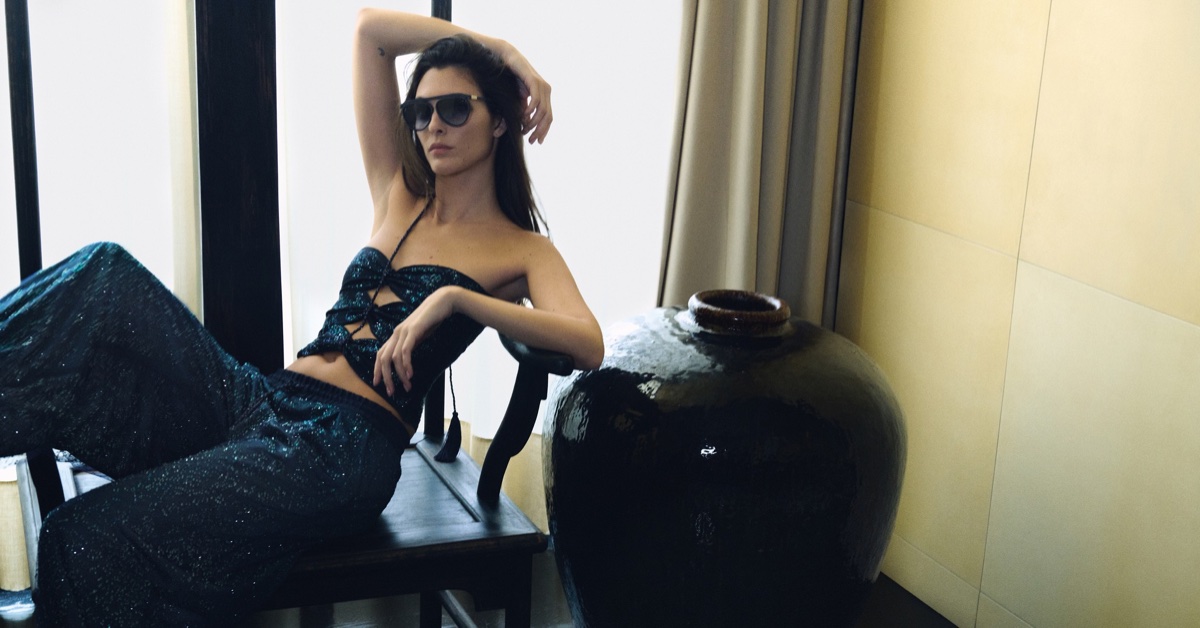 Vittoria Ceretti wearing a dark blue sparkling top and matching trousers, leaning on a dark wooden chair near a large ceramic vase.