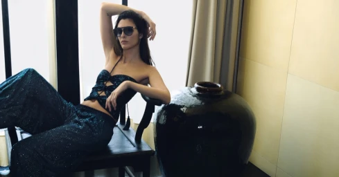 Vittoria Ceretti wearing a dark blue sparkling top and matching trousers, leaning on a dark wooden chair near a large ceramic vase.