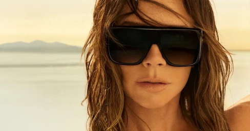 Victoria Beckham wearing oversized black sunglasses against a hazy coastal sunset background