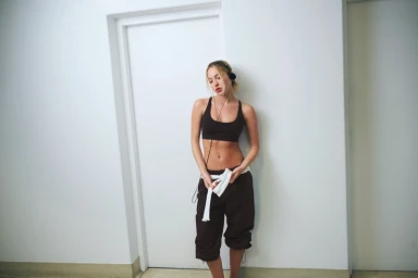 Model Lila Moss wearing a brown athletic crop top and trousers with a white tie against a plain wall for Adanola.