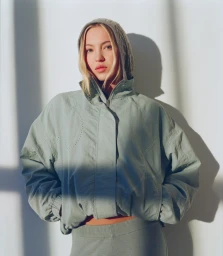 Lila Moss in a green hooded jacket and matching bottoms against a white wall with strong window light and shadow casting.