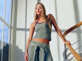 Model Lila Moss in sage green Adanola sportswear leaning on a curved wooden stair rail indoors by windows