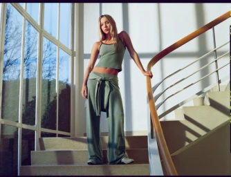 Model Lila Moss standing on indoor stairs wearing a green ribbed tank top and matching trousers for Adanola.