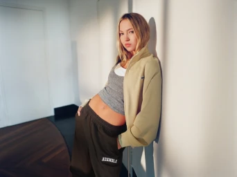 Model Lila Moss leaning against a white wall wearing a beige zip jacket, grey tank top, and dark track pants for Adanola.