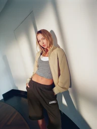 Model Lila Moss wearing a cream zip-up jacket, grey crop top, and dark trousers against a white wall.