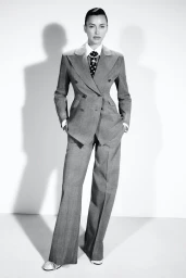 Model Irina Shayk wearing a tailored grey double-breasted suit and polka dot tie against a seamless white background.