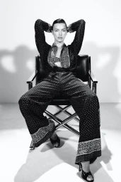 Model Irina Shayk seated in a black leather chair wearing patterned trousers and a matching long-sleeve top against a white backdrop.