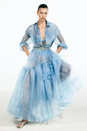 Irina Shayk wearing a sheer light blue button-down shirt and layered tulle maxi skirt against a white studio background.