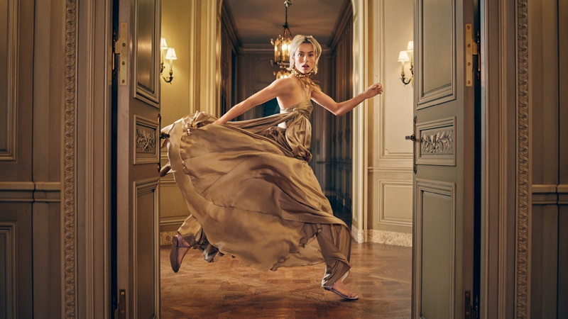 Model in a flowing golden gown running through double doors in a hotel lobby for Zimmermann Spring 2026 campaign.