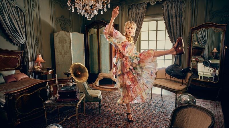 Model in a colourful floral tiered dress lifting her leg high inside a vintage bedroom with classic wooden furniture and a chandelier.