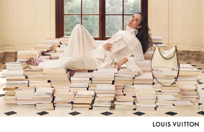 Jennifer Connelly lounging on a large pile of books holding an open book with a cream handbag nearby