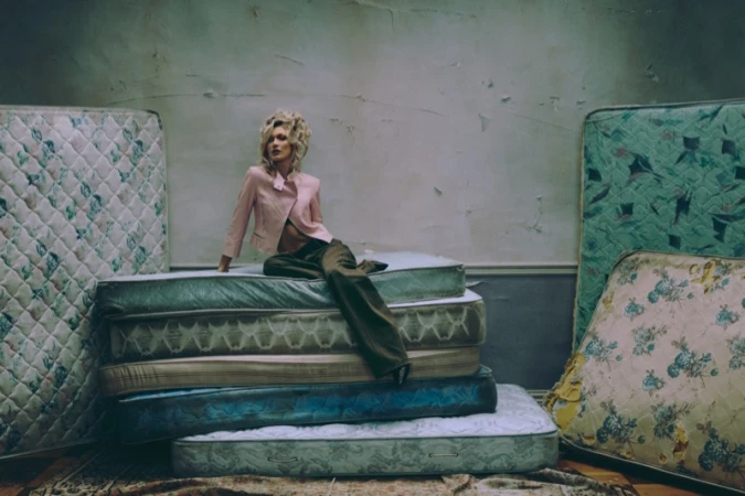 Bella Hadid poses seated on a stack of vintage mattresses wearing a cropped pink jacket and brown trousers in a distressed studio room.
