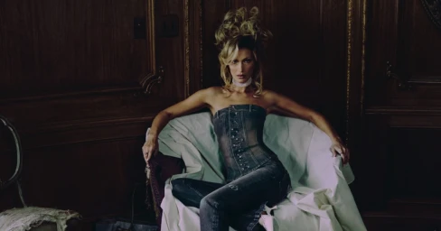 Bella Hadid in a denim outfit seated on a draped armchair against dark wood paneling for Miss Sixty