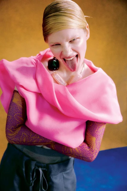 Woman with pink shawl poses with tongue out in a studio shot against a yellow background. She wears striking makeup and a large necklace.