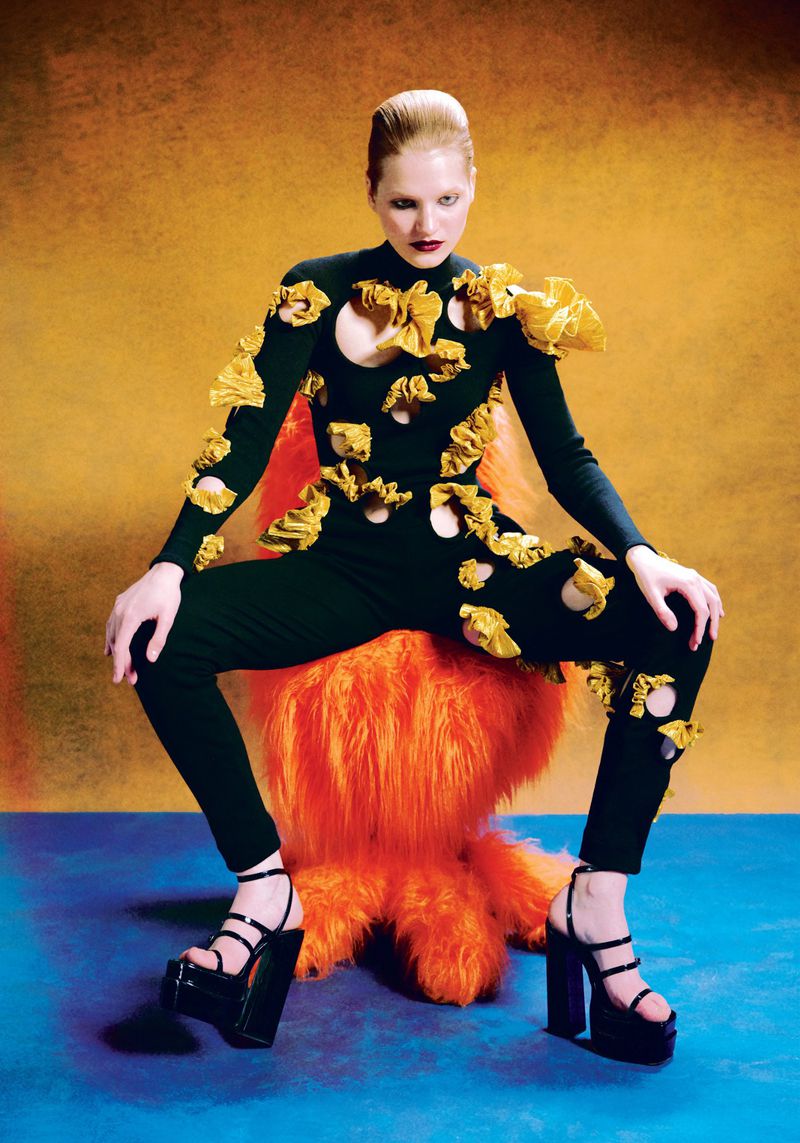 A fair-skinned adult woman in a black, sculptural jumpsuit with gold accents and platform heels sits on a furry orange chair against gradient backdrop.