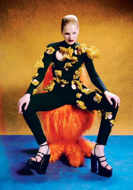 A fair-skinned adult woman in a black, sculptural jumpsuit with gold accents and platform heels sits on a furry orange chair against gradient backdrop.