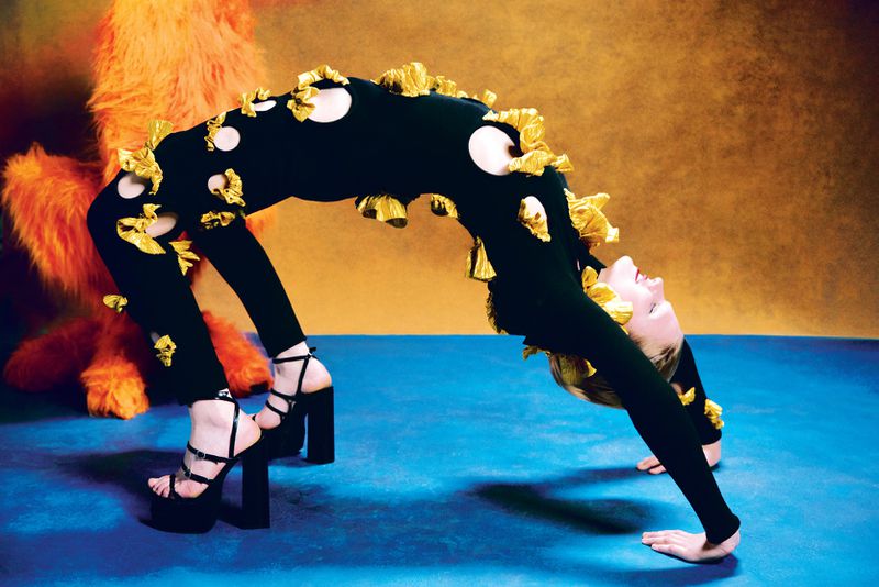 A blonde woman performs a gymnastic backbend wearing a black leotard with gold accents against a blue floor and gold backdrop.