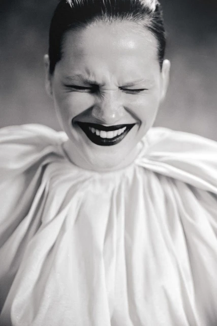 Monochrome portrait of a woman with slicked-back hair, closed eyes, dark lipstick, and wearing a large white ruffled collar.