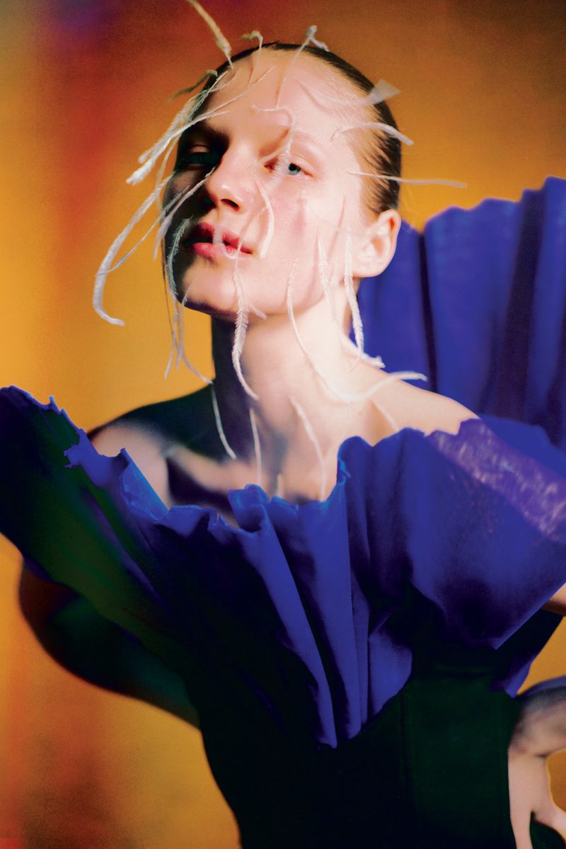 Portrait of a fair-skinned woman with white strands on her face, wearing a blue ruffled garment against an orange and red backdrop.