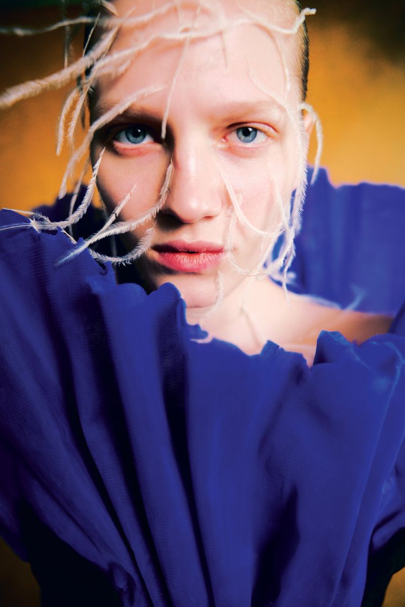 Close-up portrait of a woman with light skin and blue eyes, adorned with white feathers, wearing a blue ruffled collar against a gold background.
