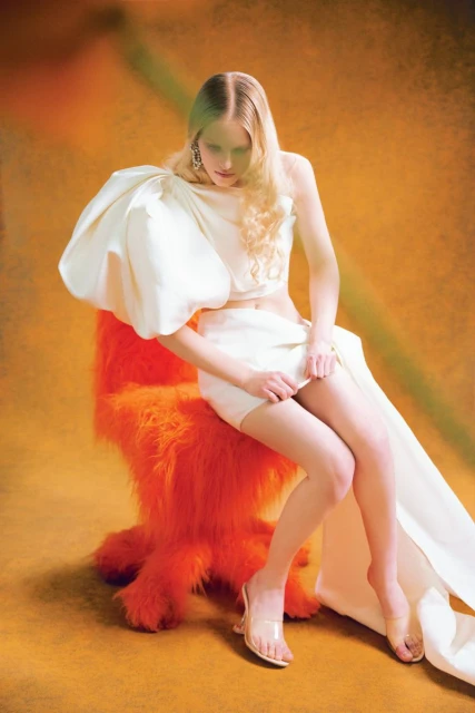 Fashionable woman in a white dress with oversized sleeve posing in a studio with an orange faux fur chair against an orange backdrop.